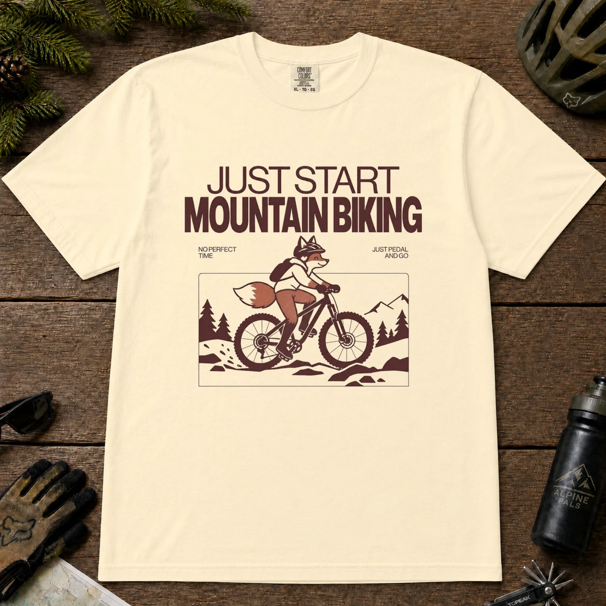 Ivory tee featuring a fox mountain biking through a minimalist trail scene with the phrases “Just Start Mountain Biking,” “No Perfect Time,” and “Just Pedal and Go.”
