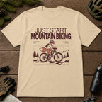 Sand tee featuring a fox mountain biking through a minimalist trail scene with the phrases “Just Start Mountain Biking,” “No Perfect Time,” and “Just Pedal and Go.”
