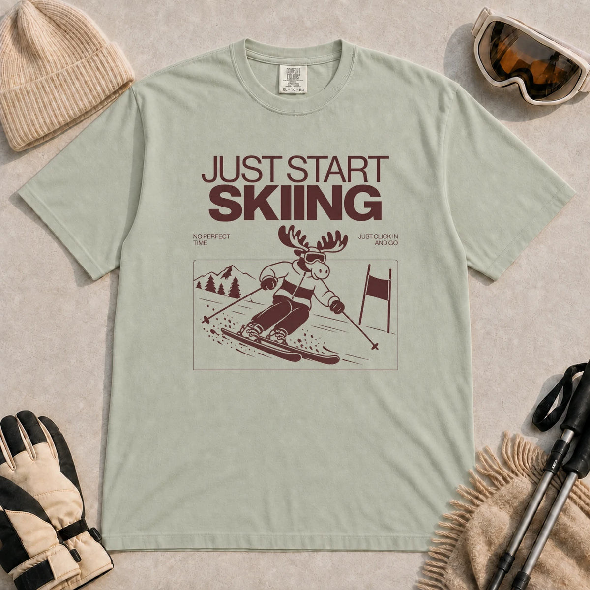 Bay tee featuring a moose skiing through a minimalist mountain scene with the phrases “Just Start Skiing,” “No Perfect Time,” and “Just Click In and Go.”