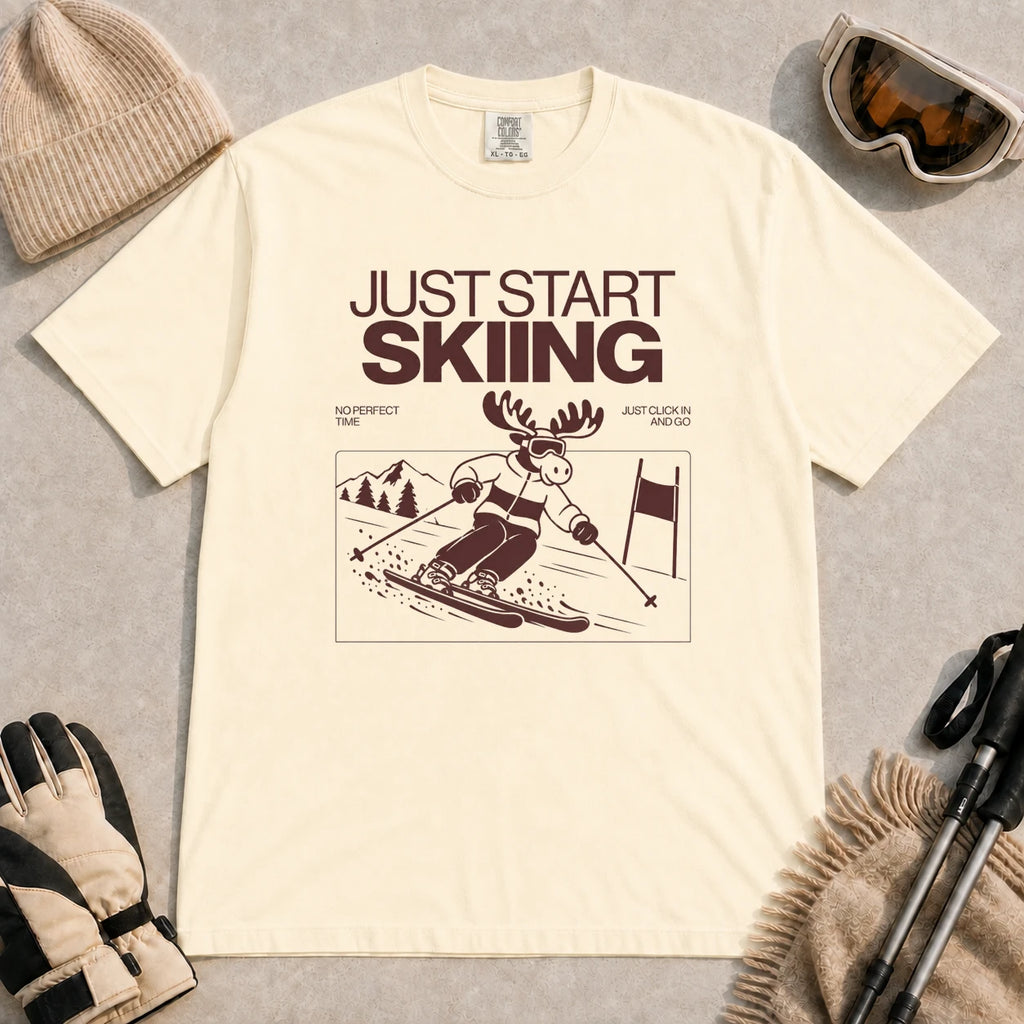 Ivory tee featuring a moose skiing through a minimalist mountain scene with the phrases “Just Start Skiing,” “No Perfect Time,” and “Just Click In and Go.”