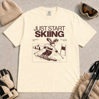 Ivory tee featuring a moose skiing through a minimalist mountain scene with the phrases “Just Start Skiing,” “No Perfect Time,” and “Just Click In and Go.”