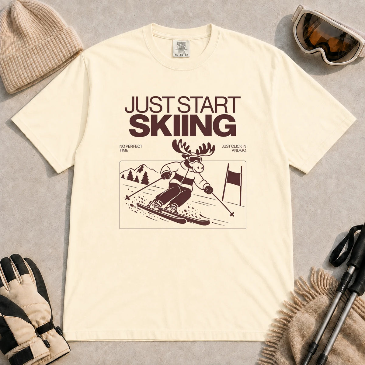 Ivory tee featuring a moose skiing through a minimalist mountain scene with the phrases “Just Start Skiing,” “No Perfect Time,” and “Just Click In and Go.”