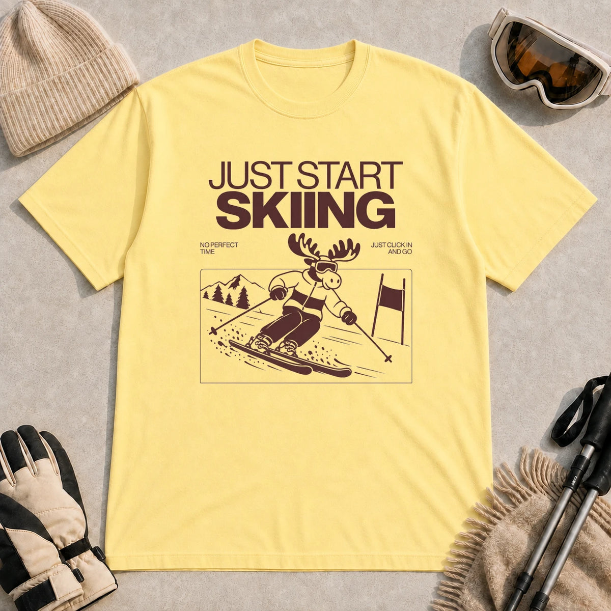Cornsilk tee featuring a moose skiing through a minimalist mountain scene with the phrases “Just Start Skiing,” “No Perfect Time,” and “Just Click In and Go.”