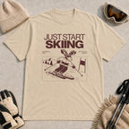 Sand tee featuring a moose skiing through a minimalist mountain scene with the phrases “Just Start Skiing,” “No Perfect Time,” and “Just Click In and Go.”