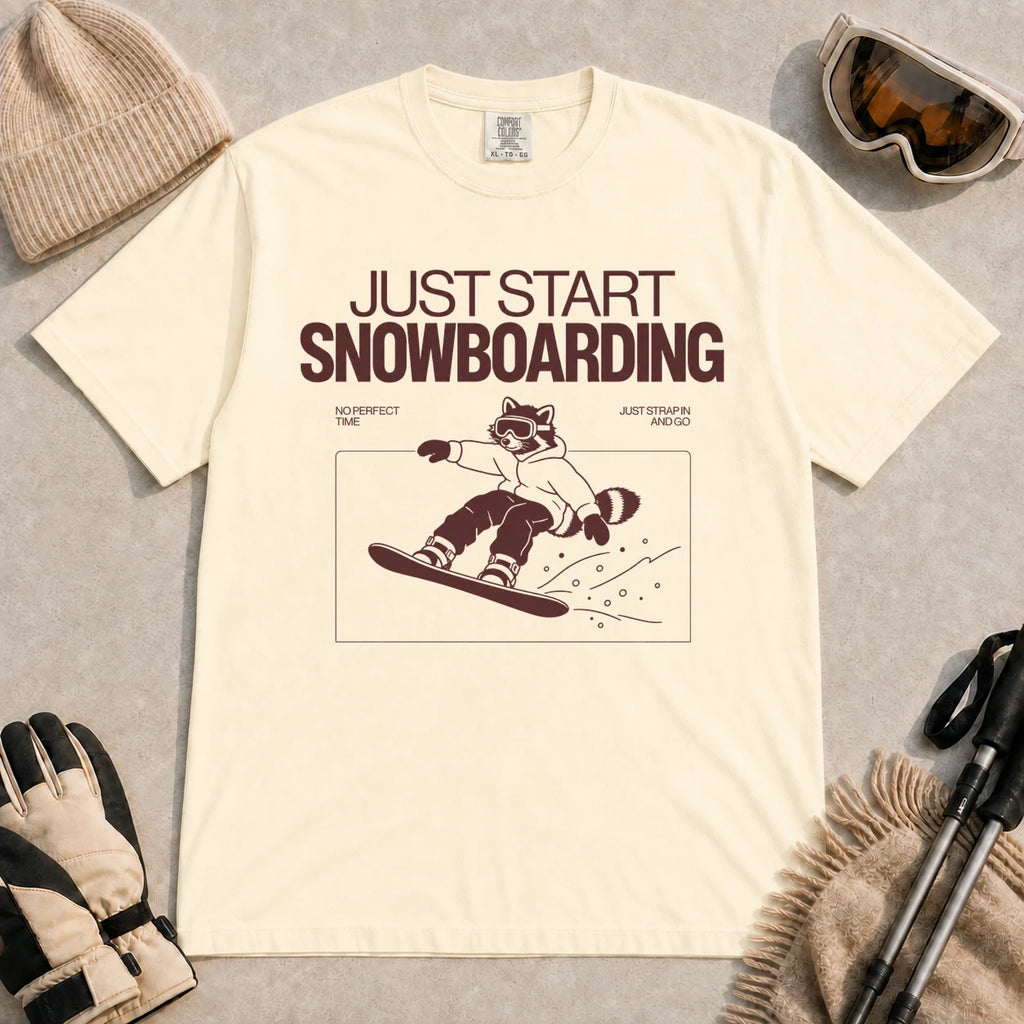 Ivory tee featuring a raccoon snowboarding through a minimalist slope scene with the phrases “Just Start Snowboarding,” “No Perfect Time,” and “Just Strap In and Go.”