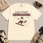 Ivory tee featuring a raccoon snowboarding through a minimalist slope scene with the phrases “Just Start Snowboarding,” “No Perfect Time,” and “Just Strap In and Go.”