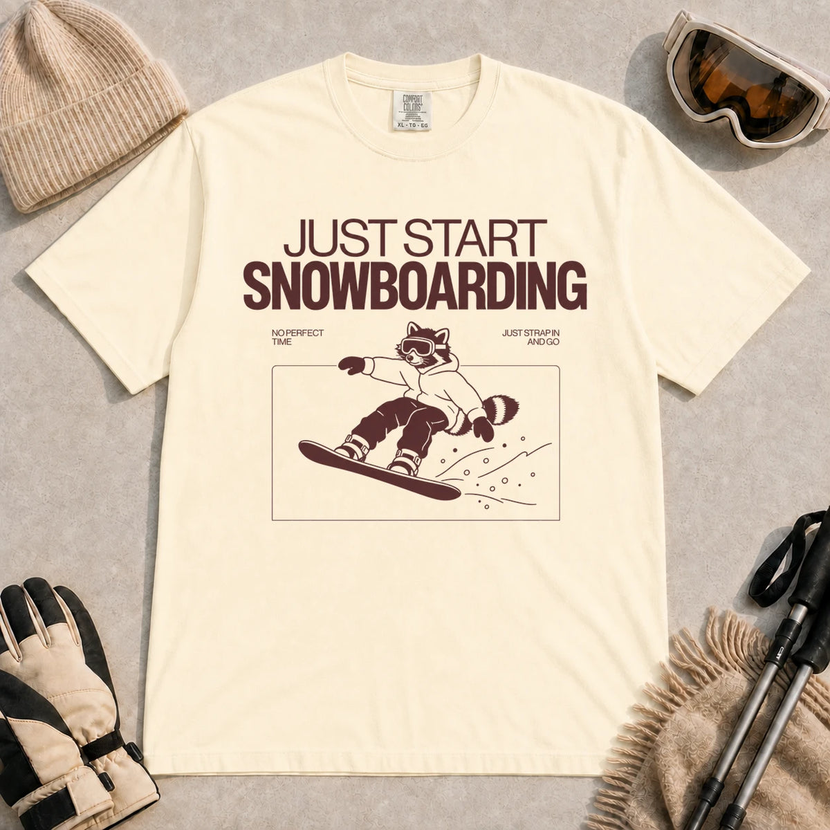 Ivory tee featuring a raccoon snowboarding through a minimalist slope scene with the phrases “Just Start Snowboarding,” “No Perfect Time,” and “Just Strap In and Go.”