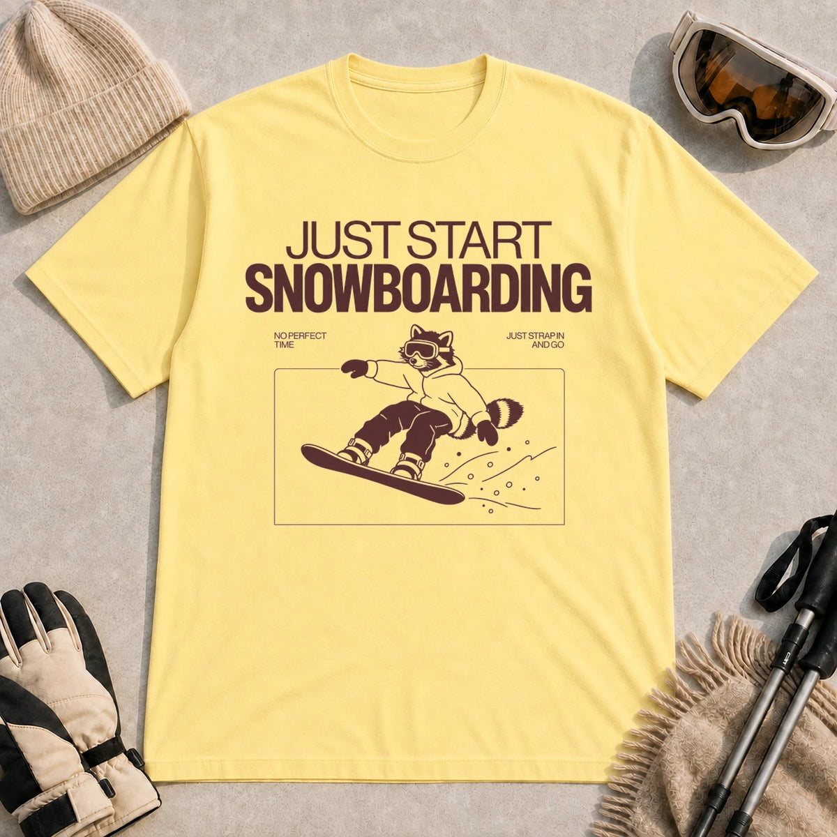 Cornsilk tee featuring a raccoon snowboarding through a minimalist slope scene with the phrases “Just Start Snowboarding,” “No Perfect Time,” and “Just Strap In and Go.”