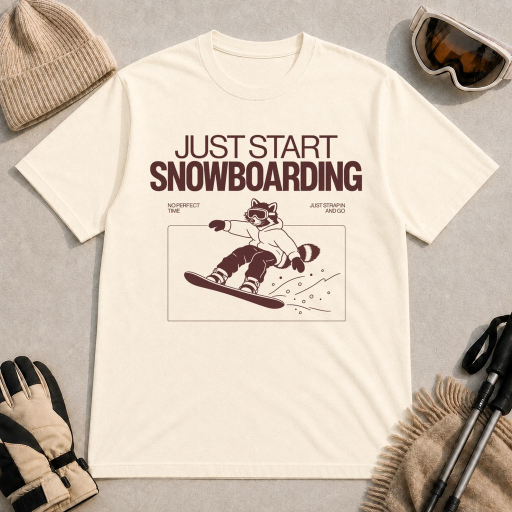 Ivory tee featuring a raccoon snowboarding through a minimalist slope scene with the phrases “Just Start Snowboarding,” “No Perfect Time,” and “Just Strap In and Go.”