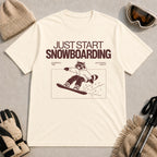 Ivory tee featuring a raccoon snowboarding through a minimalist slope scene with the phrases “Just Start Snowboarding,” “No Perfect Time,” and “Just Strap In and Go.”