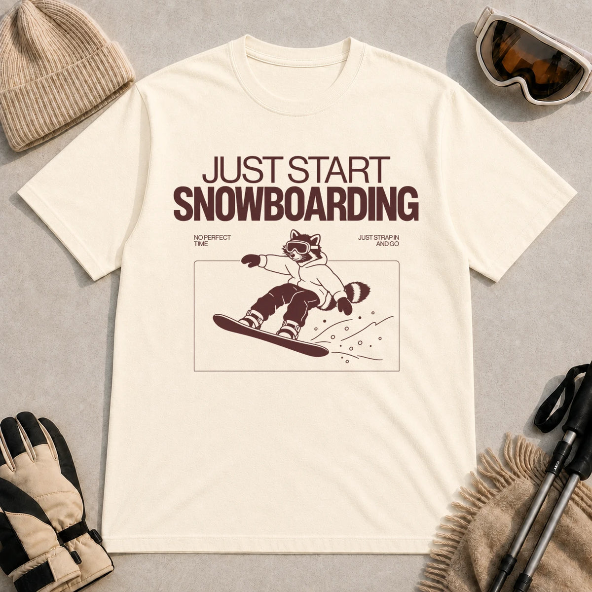 Ivory tee featuring a raccoon snowboarding through a minimalist slope scene with the phrases “Just Start Snowboarding,” “No Perfect Time,” and “Just Strap In and Go.”