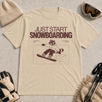 Sand tee featuring a raccoon snowboarding through a minimalist slope scene with the phrases “Just Start Snowboarding,” “No Perfect Time,” and “Just Strap In and Go.”