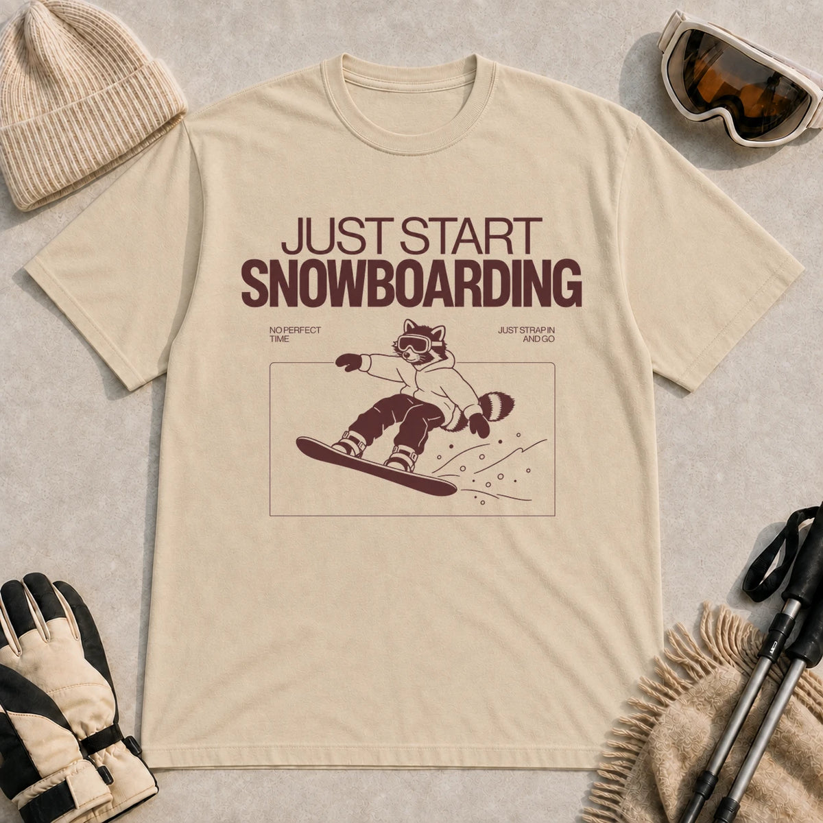Sand tee featuring a raccoon snowboarding through a minimalist slope scene with the phrases “Just Start Snowboarding,” “No Perfect Time,” and “Just Strap In and Go.”
