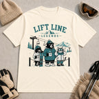 Natural tee featuring a dog, bear, and cat waiting in a ski lift line with warm drinks and winter gear, with the phrase “Lift Line Legends.”