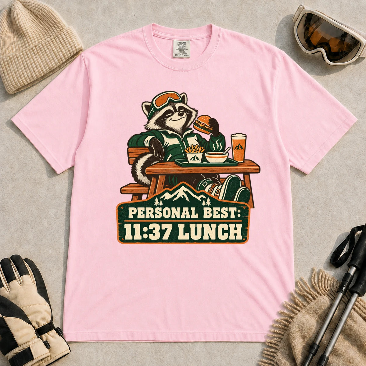 Blossom tee featuring a relaxed raccoon enjoying a burger and fries at a mountain lodge table above the phrase “Personal Best: 11:37 Lunch.”