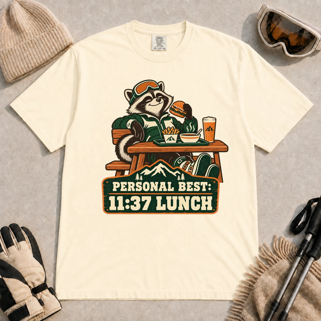 Ivory tee featuring a relaxed raccoon enjoying a burger and fries at a mountain lodge table above the phrase “Personal Best: 11:37 Lunch.”