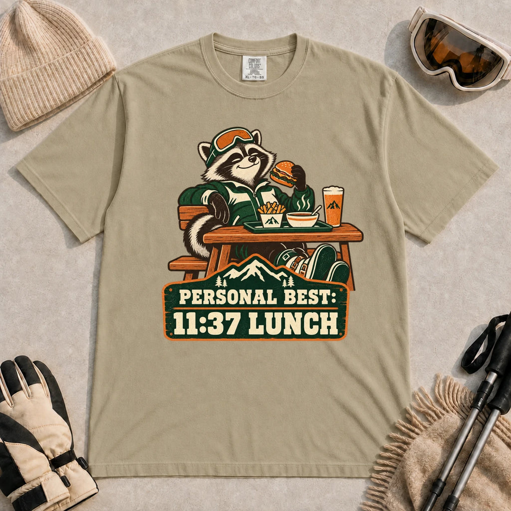 Khaki tee featuring a relaxed raccoon enjoying a burger and fries at a mountain lodge table above the phrase “Personal Best: 11:37 Lunch.”