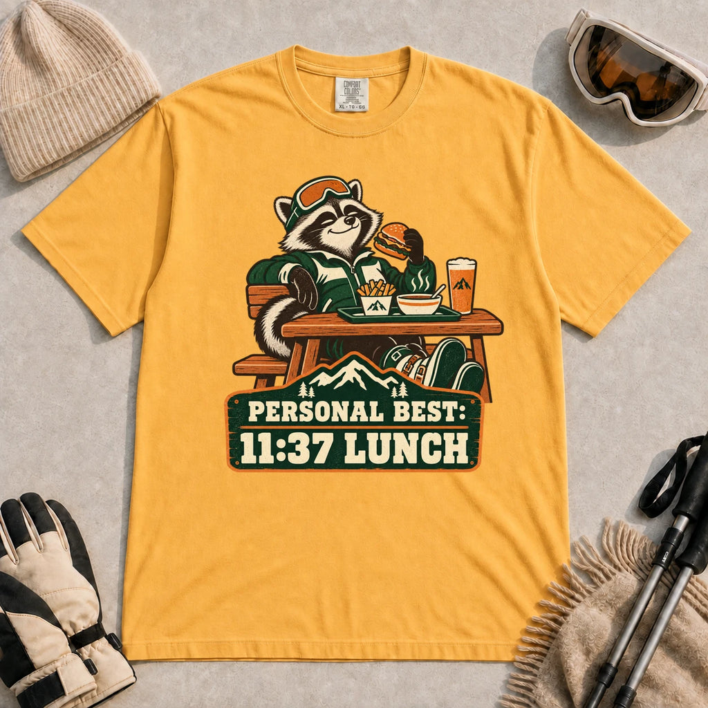 Mustard tee featuring a relaxed raccoon enjoying a burger and fries at a mountain lodge table above the phrase “Personal Best: 11:37 Lunch.”