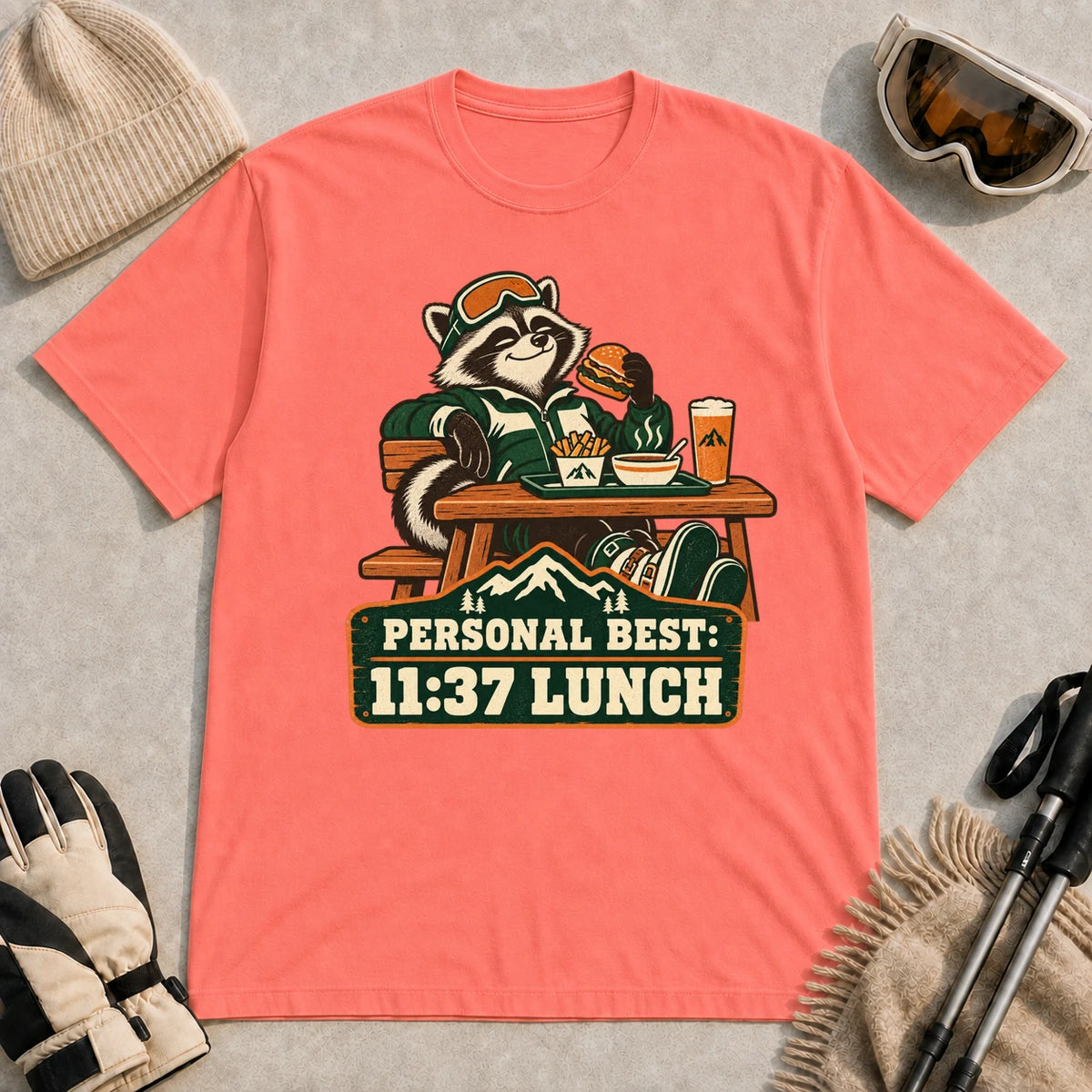Coral Silk tee featuring a relaxed raccoon enjoying a burger and fries at a mountain lodge table above the phrase “Personal Best: 11:37 Lunch.”