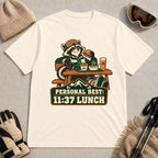 Natural tee featuring a relaxed raccoon enjoying a burger and fries at a mountain lodge table above the phrase “Personal Best: 11:37 Lunch.”