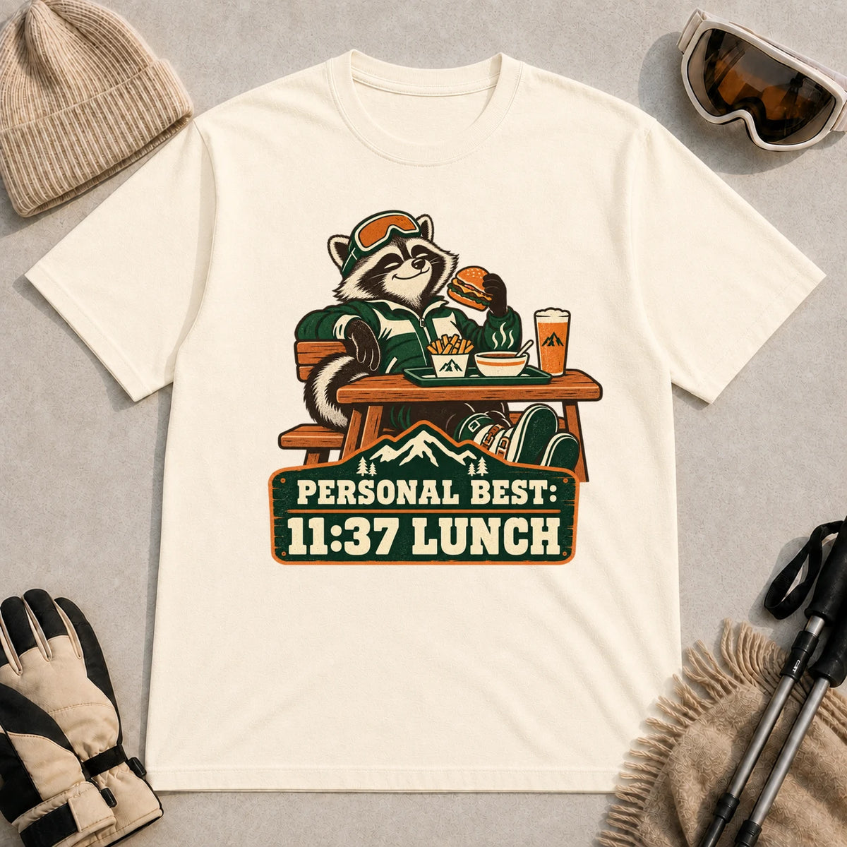 Natural tee featuring a relaxed raccoon enjoying a burger and fries at a mountain lodge table above the phrase “Personal Best: 11:37 Lunch.”