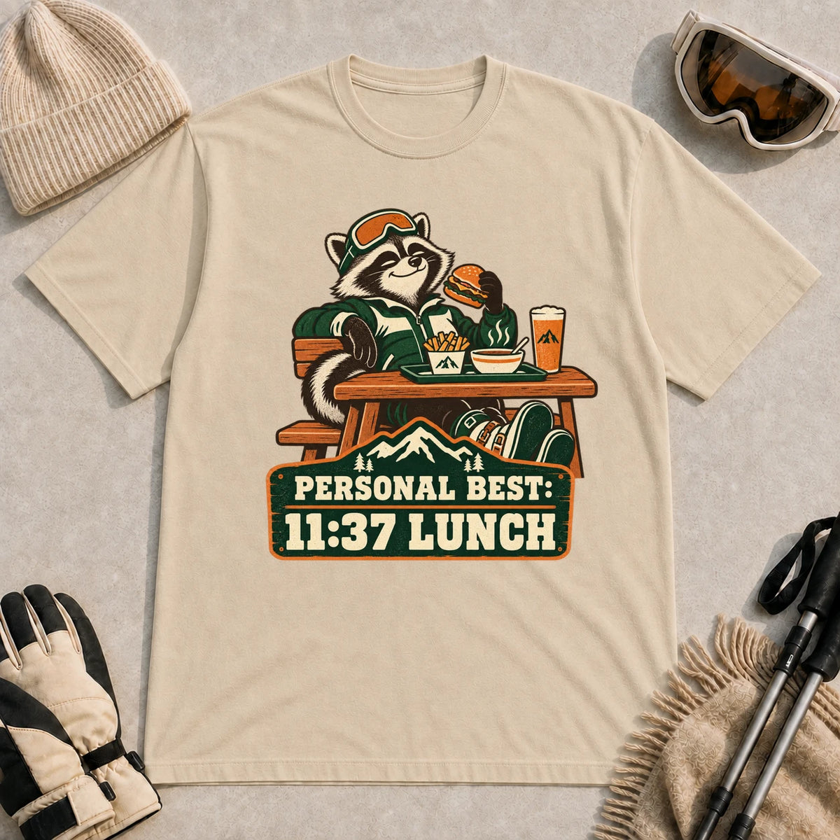 Sand tee featuring a relaxed raccoon enjoying a burger and fries at a mountain lodge table above the phrase “Personal Best: 11:37 Lunch.”