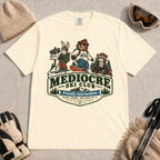 Ivory tee featuring a bear, bunny, and raccoon in ski gear above a retro “Mediocre Ski Club” badge with the phrases “Proudly Intermediate” and “Blues, Brews, and Back by 4.”