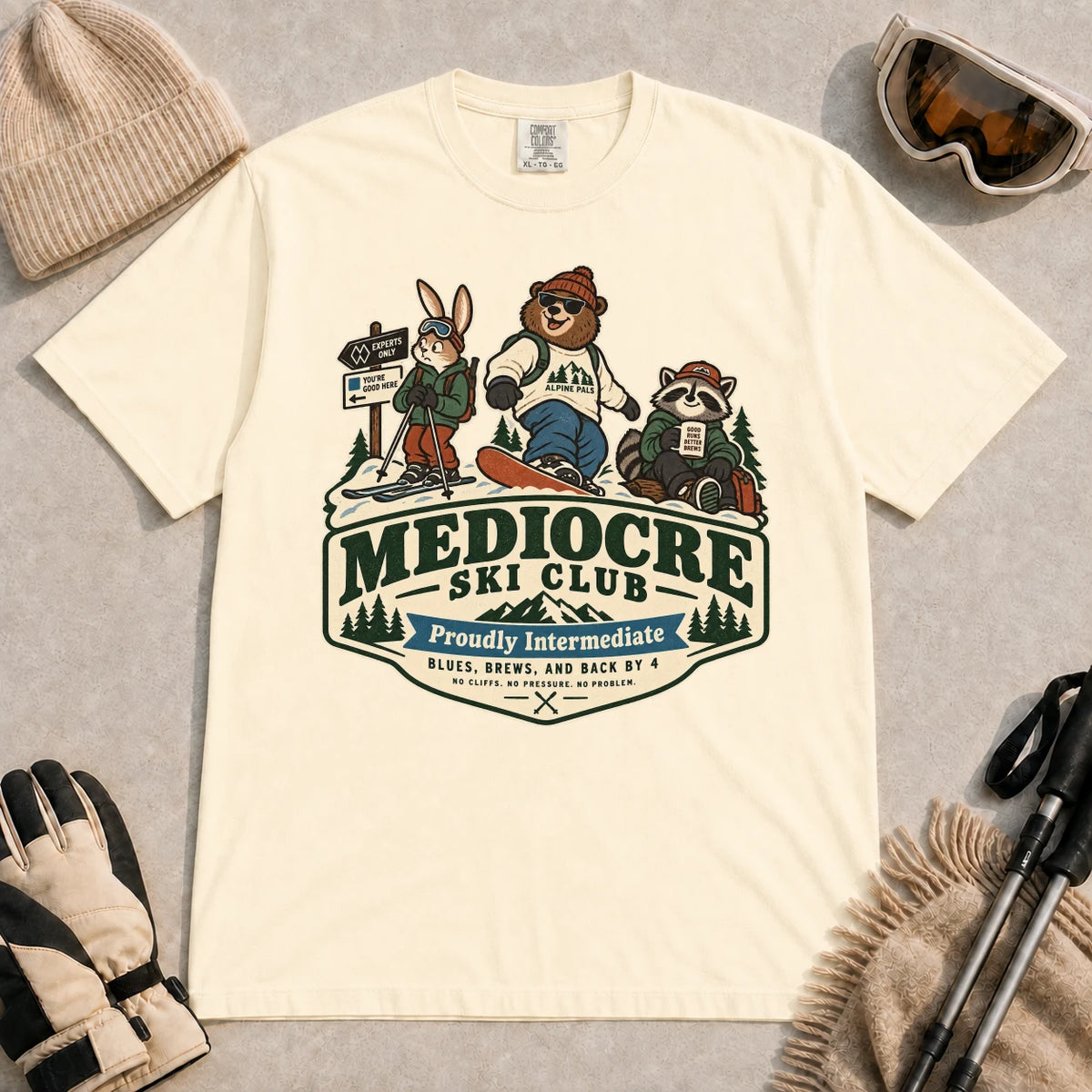 Ivory tee featuring a bear, bunny, and raccoon in ski gear above a retro “Mediocre Ski Club” badge with the phrases “Proudly Intermediate” and “Blues, Brews, and Back by 4.”