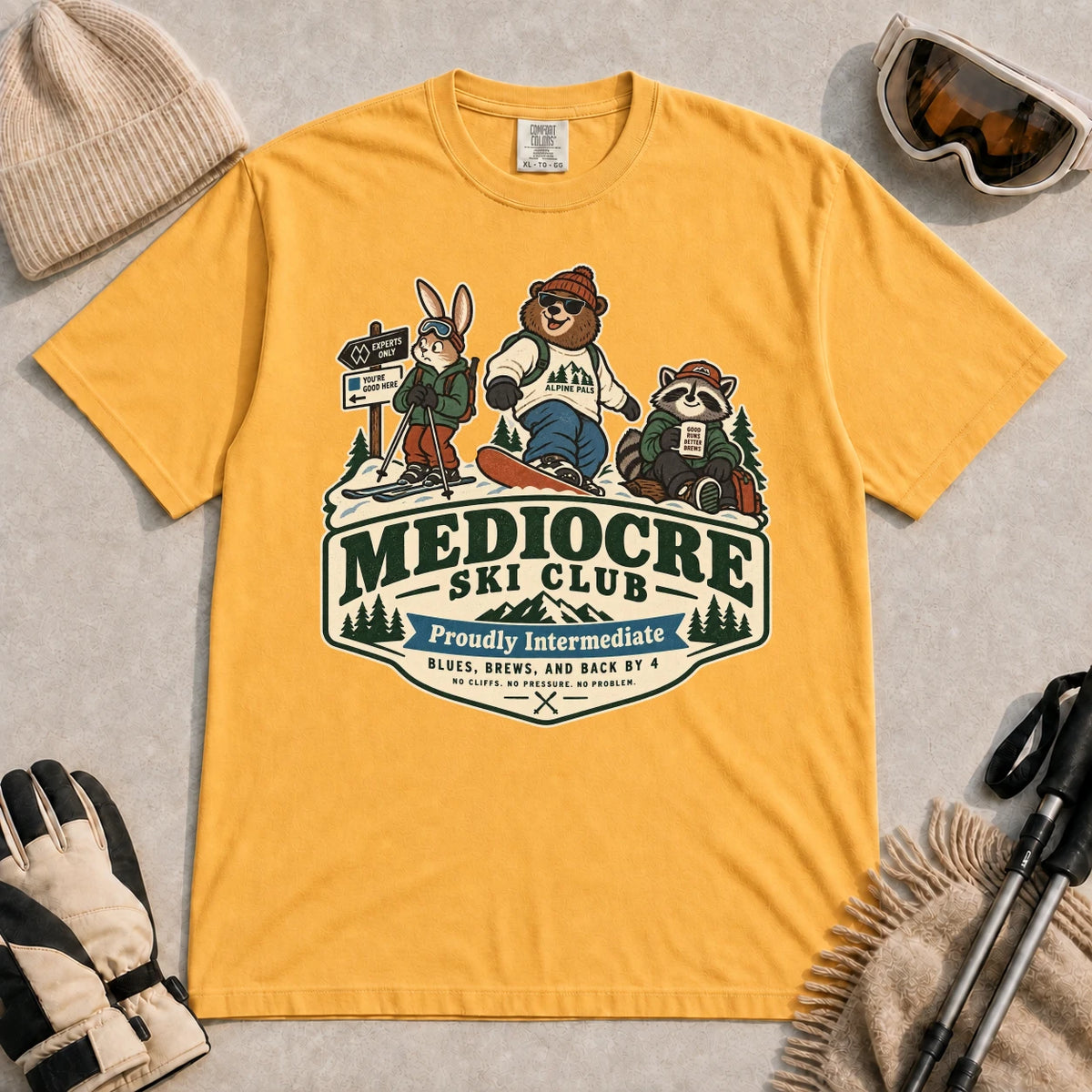 Mustard tee featuring a bear, bunny, and raccoon in ski gear above a retro “Mediocre Ski Club” badge with the phrases “Proudly Intermediate” and “Blues, Brews, and Back by 4.”