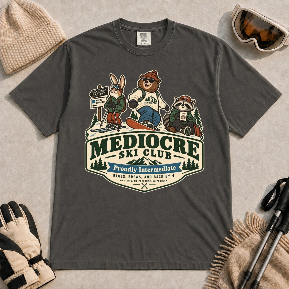 Pepper tee featuring a bear, bunny, and raccoon in ski gear above a retro “Mediocre Ski Club” badge with the phrases “Proudly Intermediate” and “Blues, Brews, and Back by 4.”