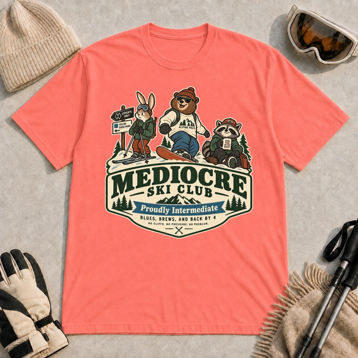Coral Silk tee featuring a bear, bunny, and raccoon in ski gear above a retro “Mediocre Ski Club” badge with the phrases “Proudly Intermediate” and “Blues, Brews, and Back by 4.”