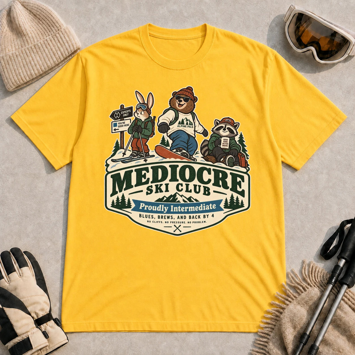Daisy tee featuring a bear, bunny, and raccoon in ski gear above a retro “Mediocre Ski Club” badge with the phrases “Proudly Intermediate” and “Blues, Brews, and Back by 4.”