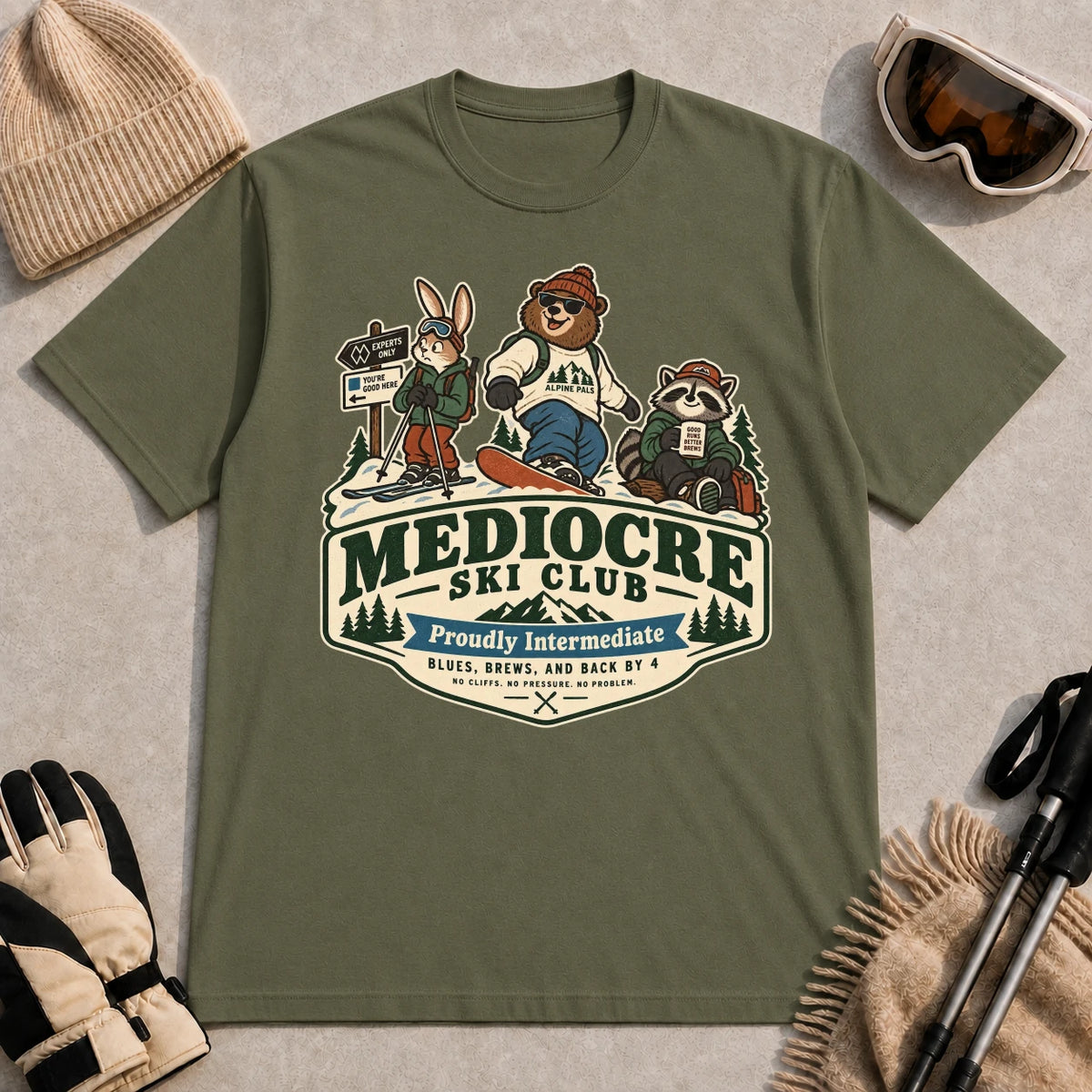 Military Green tee featuring a bear, bunny, and raccoon in ski gear above a retro “Mediocre Ski Club” badge with the phrases “Proudly Intermediate” and “Blues, Brews, and Back by 4.”