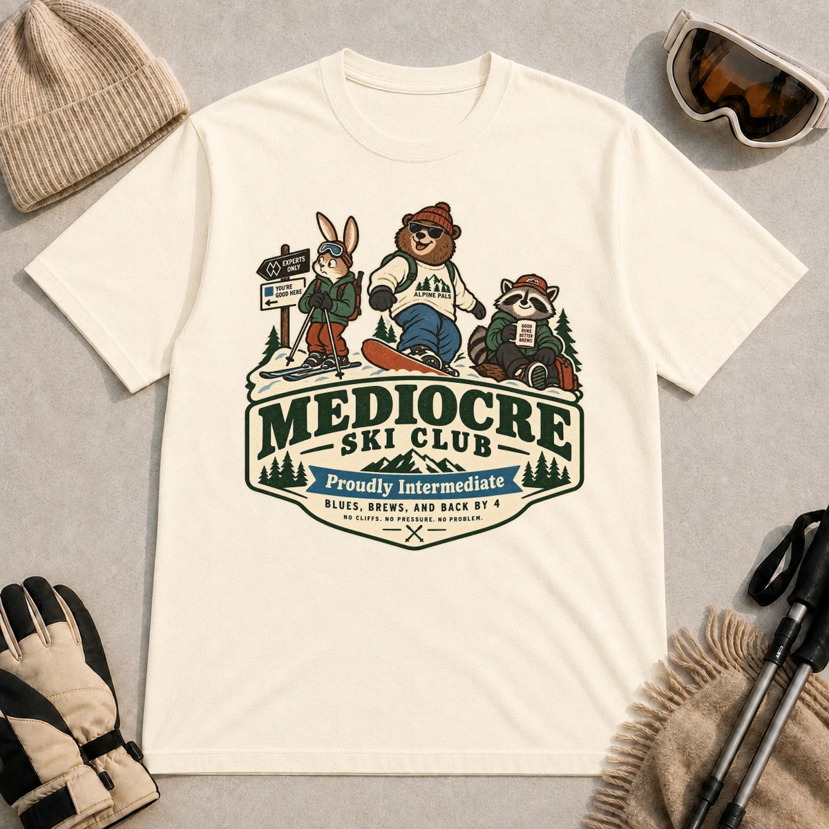 Natural tee featuring a bear, bunny, and raccoon in ski gear above a retro “Mediocre Ski Club” badge with the phrases “Proudly Intermediate” and “Blues, Brews, and Back by 4.”
