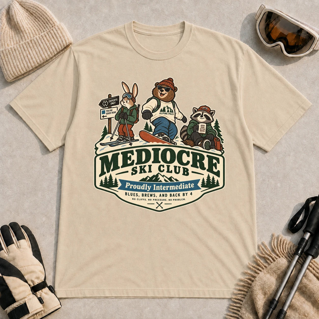 Sand tee featuring a bear, bunny, and raccoon in ski gear above a retro “Mediocre Ski Club” badge with the phrases “Proudly Intermediate” and “Blues, Brews, and Back by 4.”