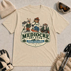 Sand tee featuring a bear, bunny, and raccoon in ski gear above a retro “Mediocre Ski Club” badge with the phrases “Proudly Intermediate” and “Blues, Brews, and Back by 4.”