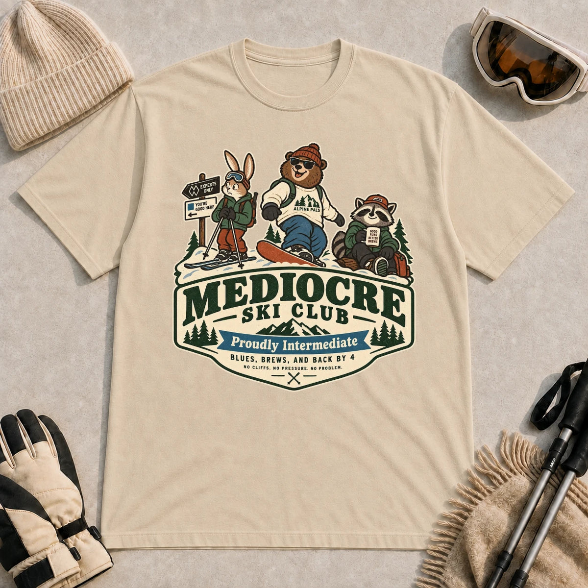 Sand tee featuring a bear, bunny, and raccoon in ski gear above a retro “Mediocre Ski Club” badge with the phrases “Proudly Intermediate” and “Blues, Brews, and Back by 4.”