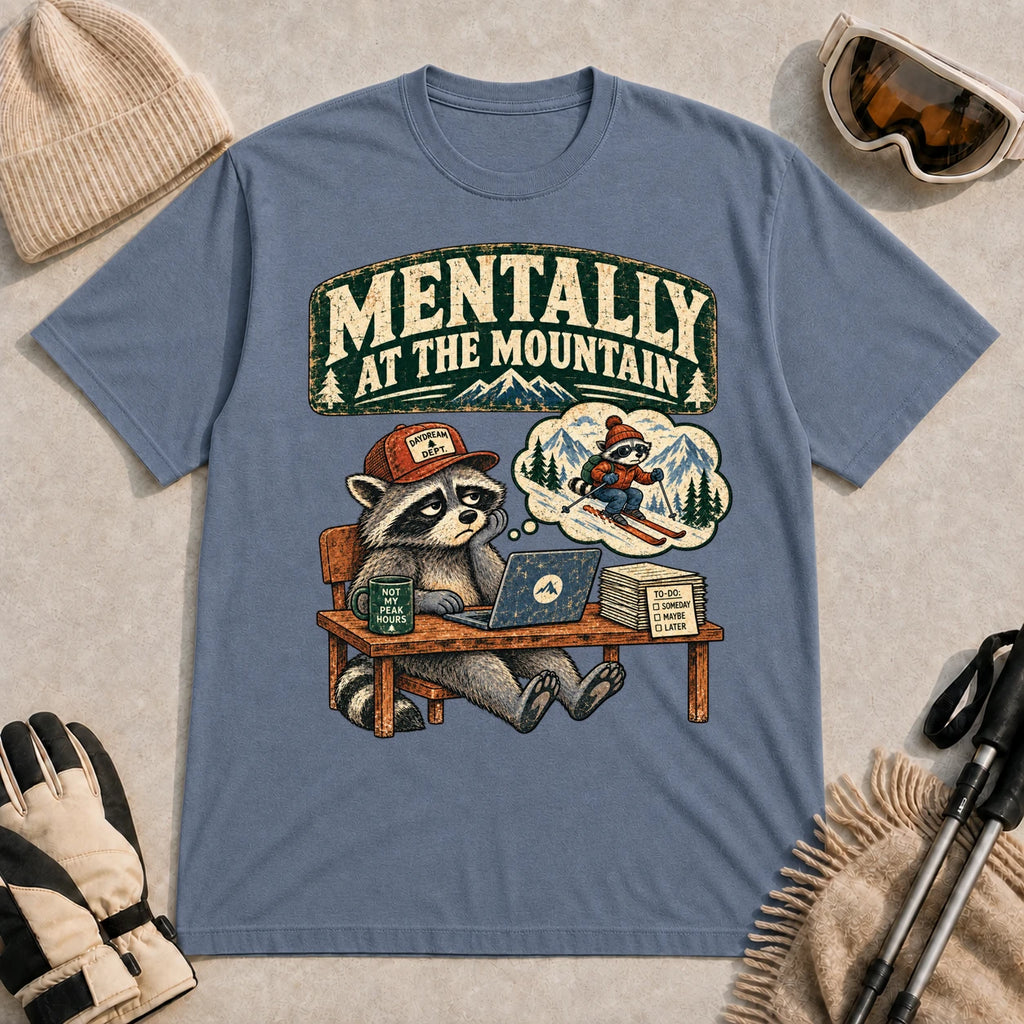 Heather Indigo tee featuring a raccoon sitting at a desk and daydreaming about skiing, with the phrase “Mentally at the Mountain.”