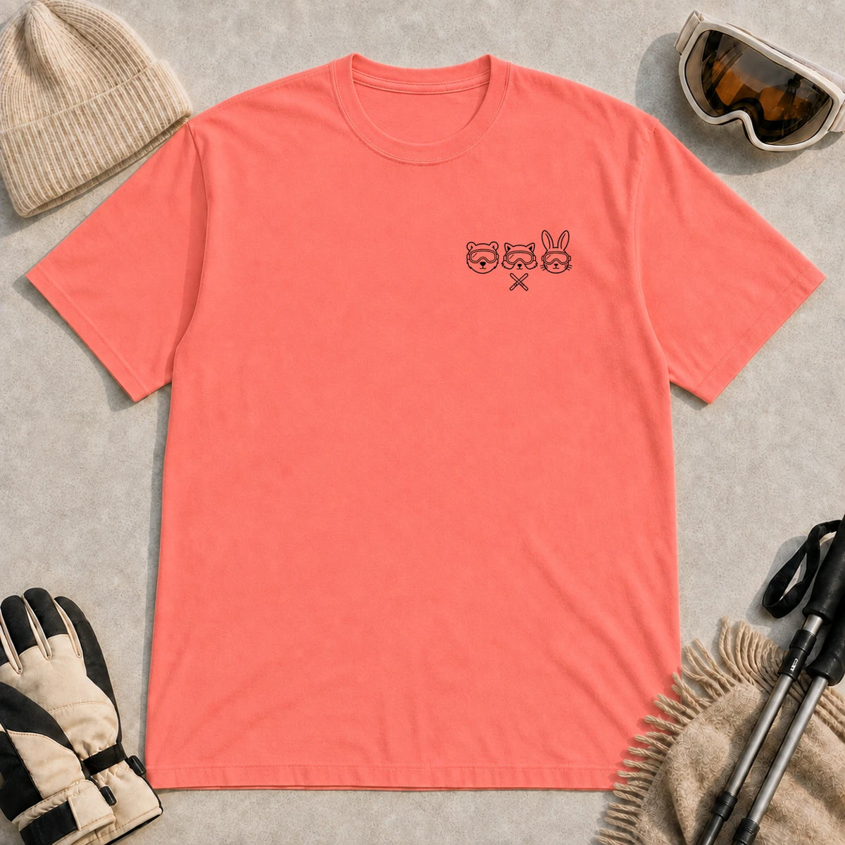 Coral Silk tee featuring minimalist black line art of a bear, raccoon, and bunny wearing ski goggles above a small pair of crossed skis.