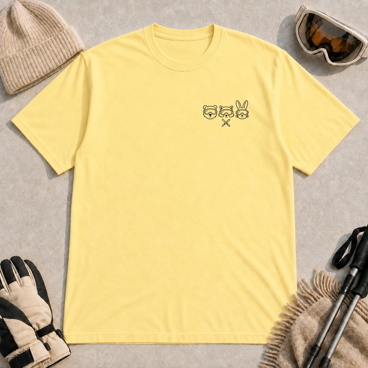 Cornsilk tee featuring minimalist black line art of a bear, raccoon, and bunny wearing ski goggles above a small pair of crossed skis.
