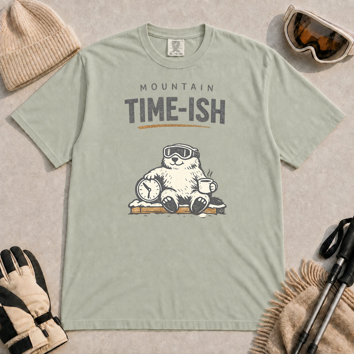 Bay tee featuring a relaxed bear in ski goggles sitting with a steaming mug and clock on a snowy bench, with the phrase “Mountain Time-ish.”