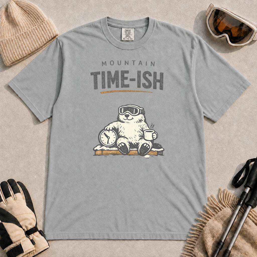 Granite tee featuring a relaxed bear in ski goggles sitting with a steaming mug and clock on a snowy bench, with the phrase “Mountain Time-ish.”