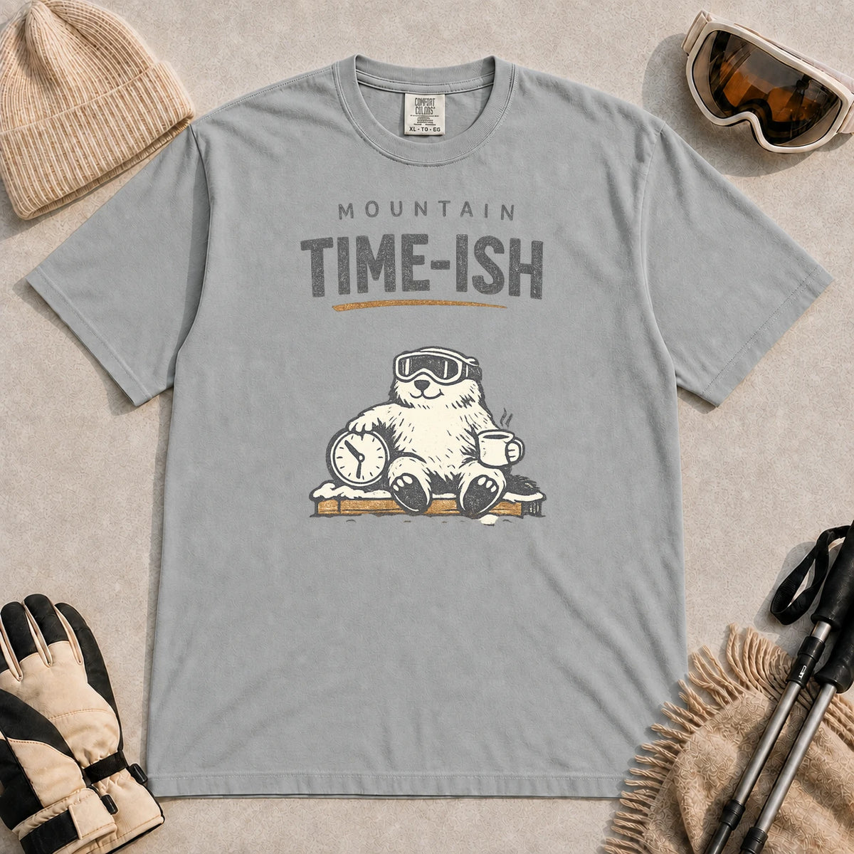Granite tee featuring a relaxed bear in ski goggles sitting with a steaming mug and clock on a snowy bench, with the phrase “Mountain Time-ish.”