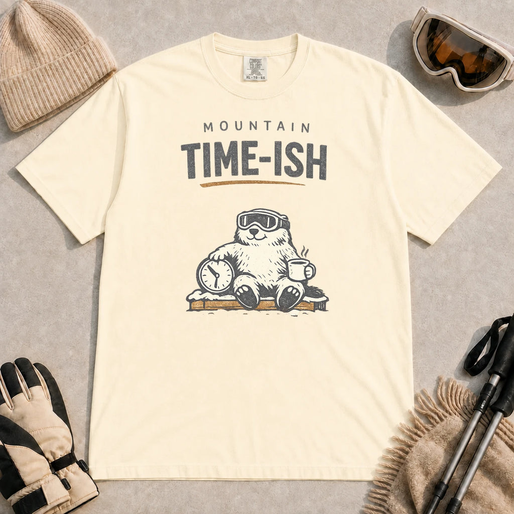 Ivory tee featuring a relaxed bear in ski goggles sitting with a steaming mug and clock on a snowy bench, with the phrase “Mountain Time-ish.”