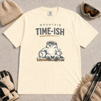 Ivory tee featuring a relaxed bear in ski goggles sitting with a steaming mug and clock on a snowy bench, with the phrase “Mountain Time-ish.”