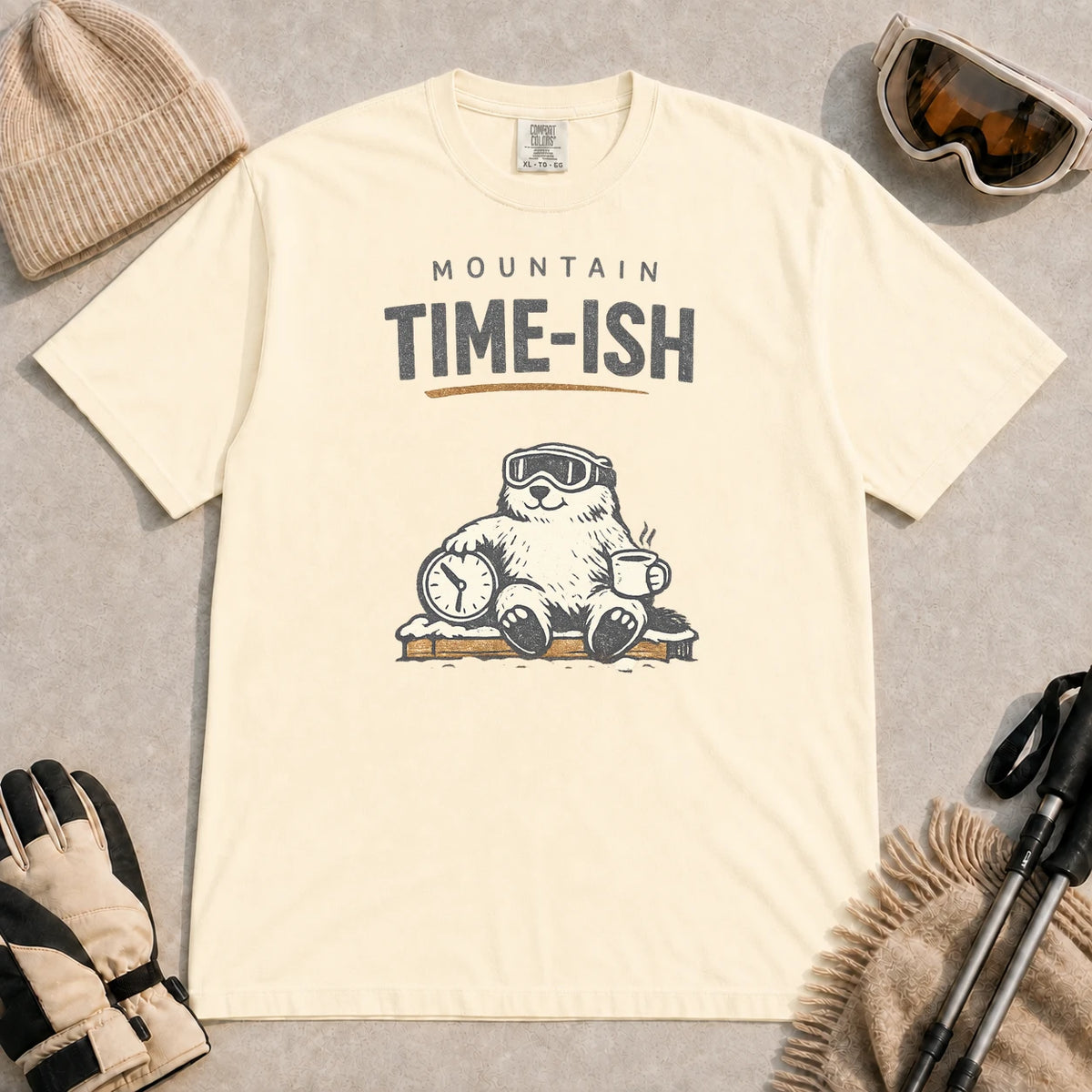 Ivory tee featuring a relaxed bear in ski goggles sitting with a steaming mug and clock on a snowy bench, with the phrase “Mountain Time-ish.”
