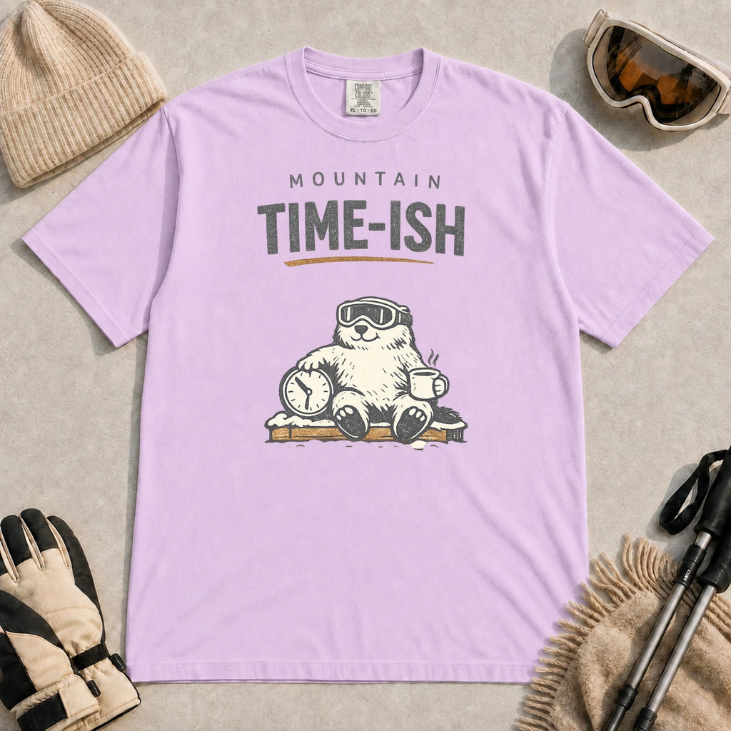 Orchid tee featuring a relaxed bear in ski goggles sitting with a steaming mug and clock on a snowy bench, with the phrase “Mountain Time-ish.”