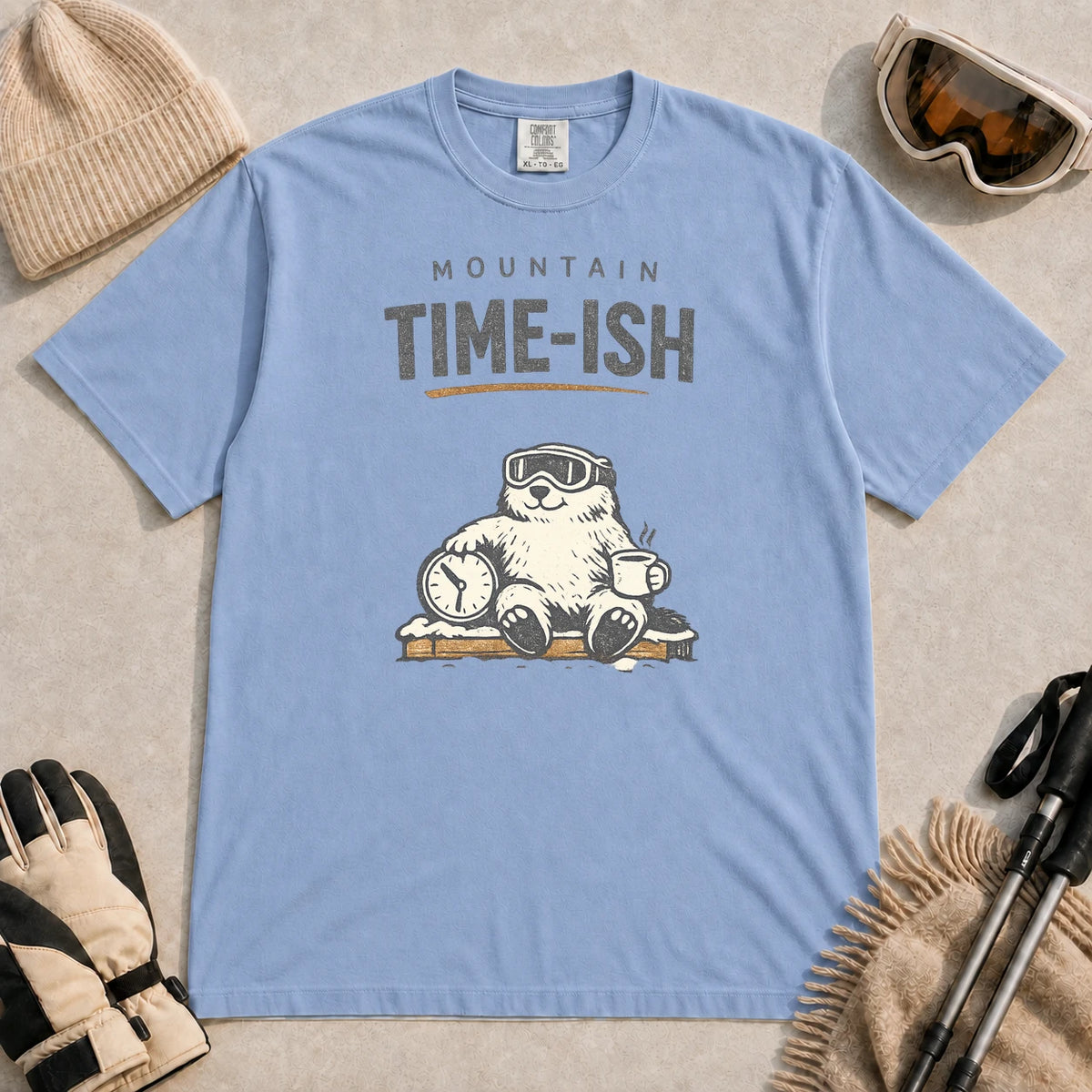 Washed Denim tee featuring a relaxed bear in ski goggles sitting with a steaming mug and clock on a snowy bench, with the phrase “Mountain Time-ish.”