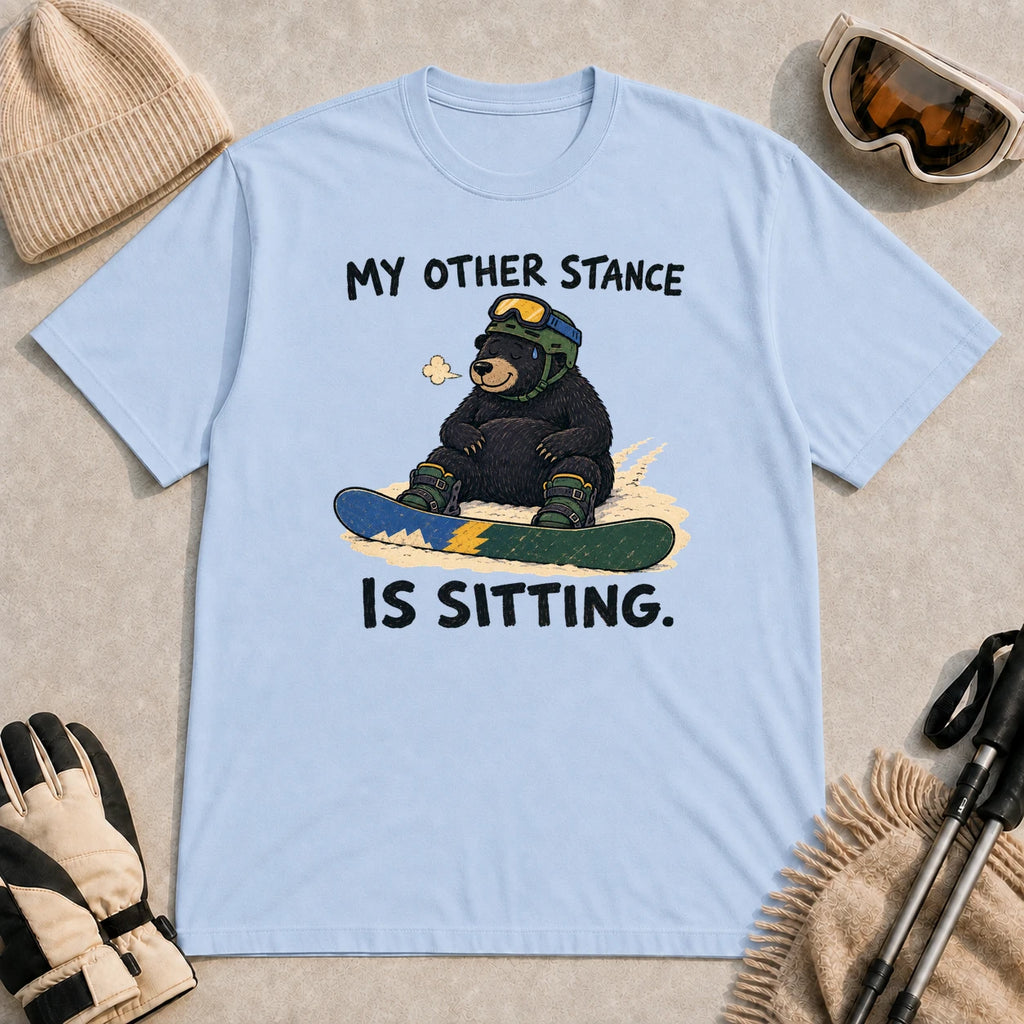 Light Blue tee featuring a black bear in snowboard gear sitting in the snow behind a snowboard with the phrase “My Other Stance Is Sitting.”