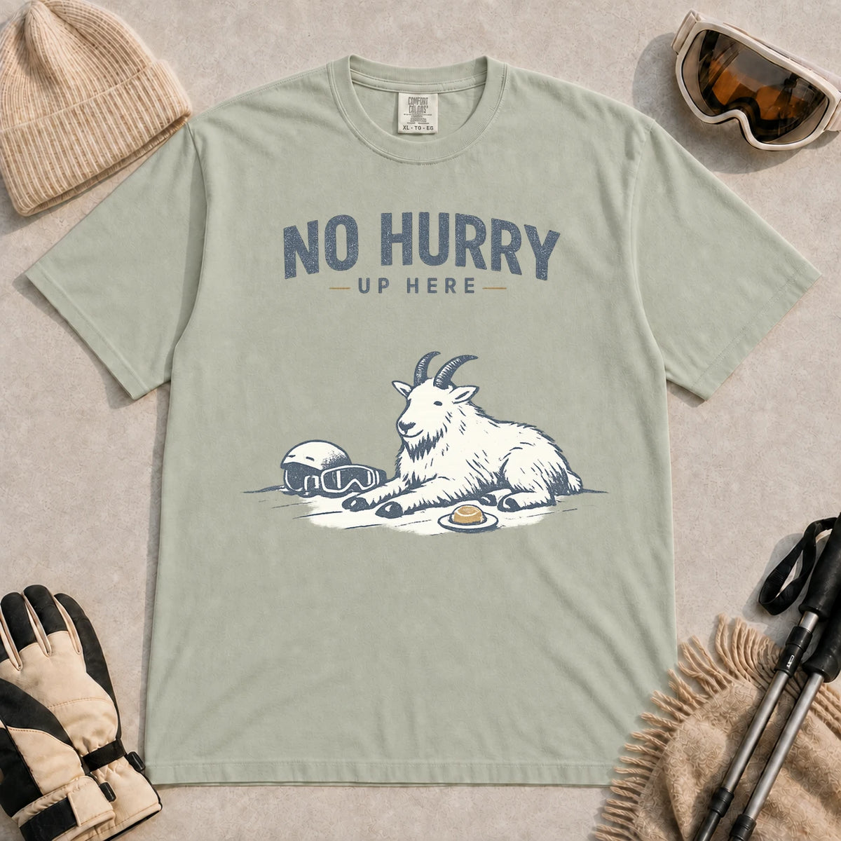 Bay tee featuring a relaxed mountain goat resting in the snow beside ski goggles and a helmet with the phrase “No Hurry Up Here.”